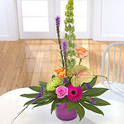 A purple vase, bouquet, with a pink rose, orange roses, a purple daisy, a Bells of Ireland flower and two purple Blazing-Stars surrounded by green leaves